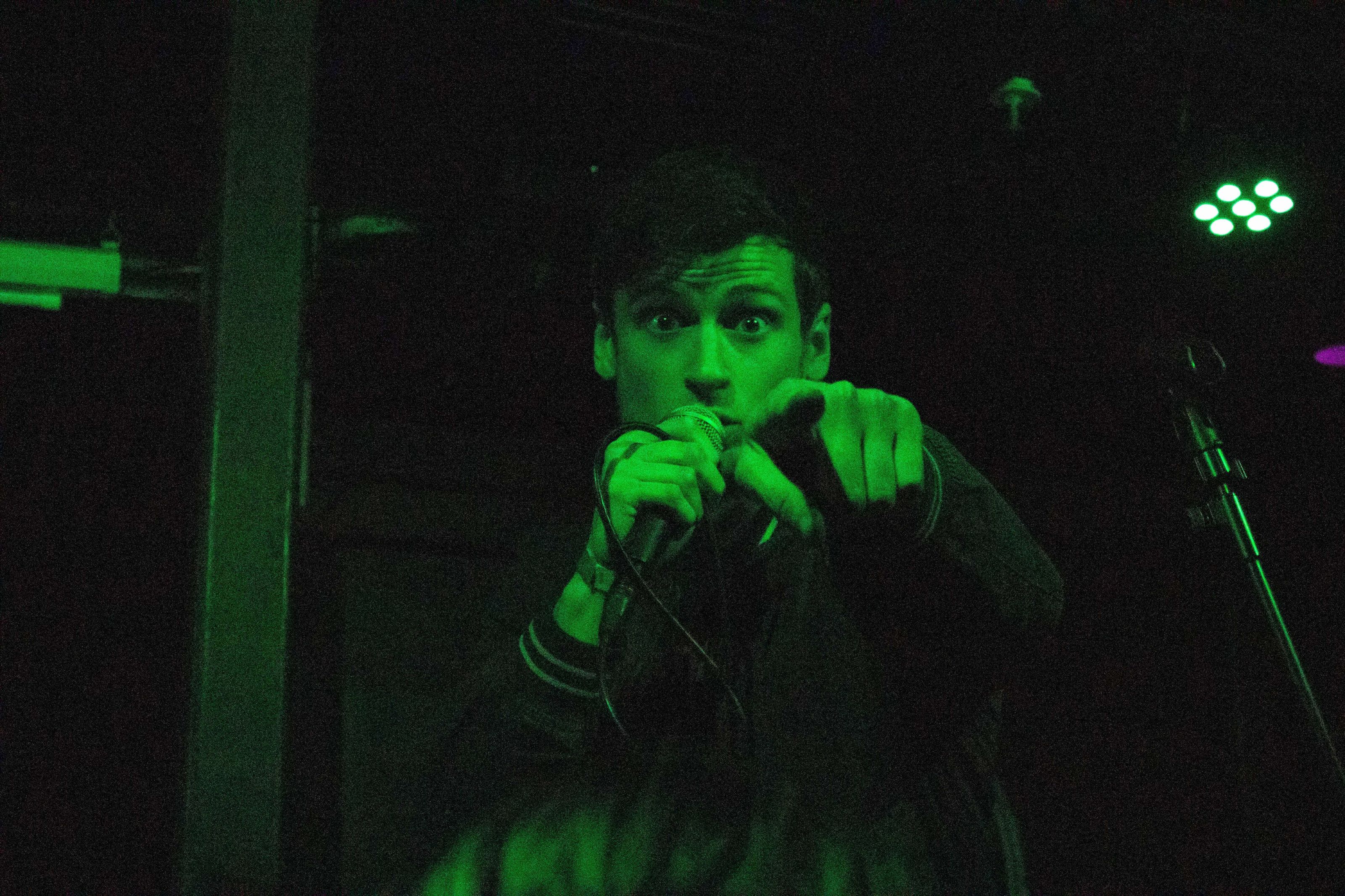 A photograph of One Trait Danger's frontman pointing at and looking directly into the camera, lit in a dark green light